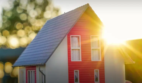 Ein Haus mit roten und weißen Wänden und einem grauen Schrägdach, beleuchtet vom warmen Schein der untergehenden Sonne im Hintergrund. Linsenreflexe und verschwommene Bäume sind zu sehen.